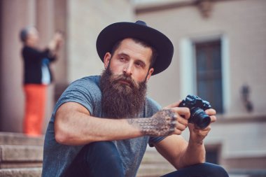 Yakışıklı hippi erkek bir şık sakal ve kollarını dövme ile rahat kıyafetler giymiş ve şapka bir kamera tutan, merdiven üstünde oturuyor.