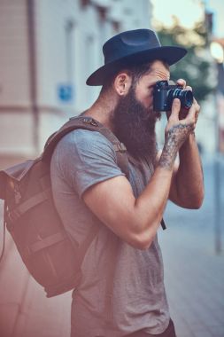 Bir fotoğraf makinesi kullanarak rahat kıyafetler içinde yakışıklı hippi erkek bir şık sakal ve kollarını dövme ile giyinmiş.