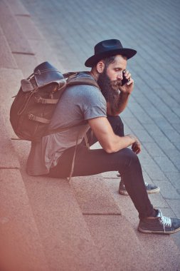 Yakışıklı hipster seyahat bir şık sakal ve kollarını dövme ile telefonda konuşurken adımlar üzerinde oturan rahat kıyafetler giymiş.