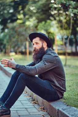 Yakışıklı hipster seyahat bir şık sakal ve kollarını rahat kıyafetler ve şapka, parkta kaldırımda oturan giymiş dövme.