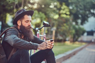 Bisiklete binmek sonra dinlenme kaldırımda oturan smartphone kullanarak yakışıklı hipster seyahat bir şık sakal ve kollarını bir çanta, şapka ve rahat kıyafetler giymiş dövme.