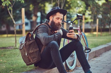 Bisiklete binmek sonra dinlenme kaldırımda oturan smartphone kullanarak yakışıklı hipster seyahat bir şık sakal ve kollarını bir çanta, şapka ve rahat kıyafetler giymiş dövme.