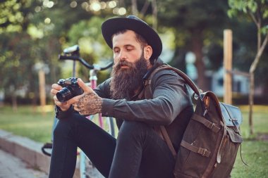 Bisiklete binmek sonra dinlenme kaldırımda oturan smartphone kullanarak yakışıklı hipster seyahat bir şık sakal ve kollarını bir çanta, şapka ve rahat kıyafetler giymiş dövme.