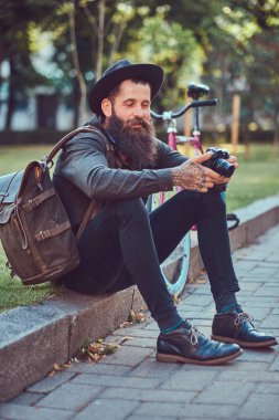 Bisiklete binmek sonra dinlenme kaldırımda oturan smartphone kullanarak yakışıklı hipster seyahat bir şık sakal ve kollarını bir çanta, şapka ve rahat kıyafetler giymiş dövme.