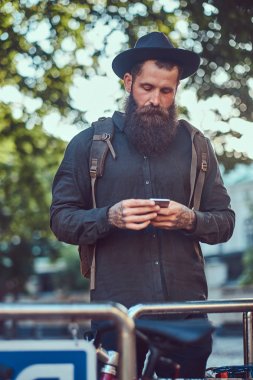 Bisiklet park yeri üzerinde duran telefon kullanarak rahat kıyafetler ve şapka, yakışıklı hipster seyahat bir şık sakal ve kollarını dövmesini giymiş.