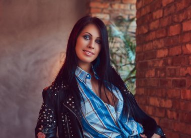 Gülümseyen bir seksi seksi esmer bir şık deri ceket ve kot bir çatı katı iç ile bir odada poz portresi.