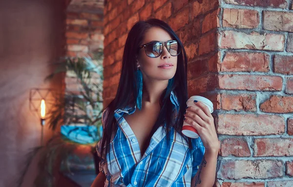 A sexy brunette in a flannel shirt and jeans holds a cup of coffee posing in a room.