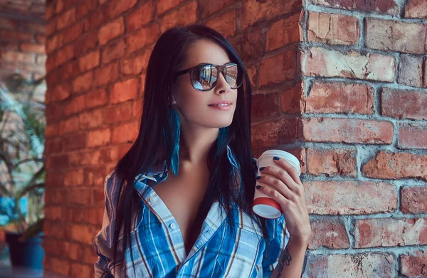 A sexy brunette in a flannel shirt and jeans holds a cup of coffee posing in a room.