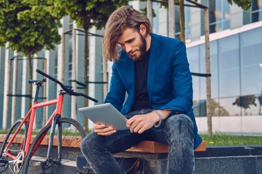 Yakışıklı şık erkek uzun saçlı bir bisiklet ile bir bankta oturmuş bir tablet bilgisayar kullanarak sakallı.