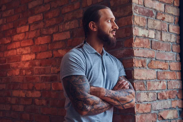 Handsome tattooed male with a stylish haircut and beard, in a gray t-shirt, standing leaning against a brick wall in a room with a loft interior.