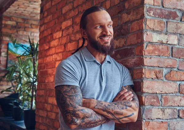 Handsome tattooed male with a stylish haircut and beard, in a gray t-shirt, standing leaning against a brick wall in a room with a loft interior.