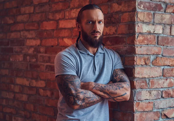 Handsome tattooed male with a stylish haircut and beard, in a gray t-shirt, standing leaning against a brick wall in a room with a loft interior.