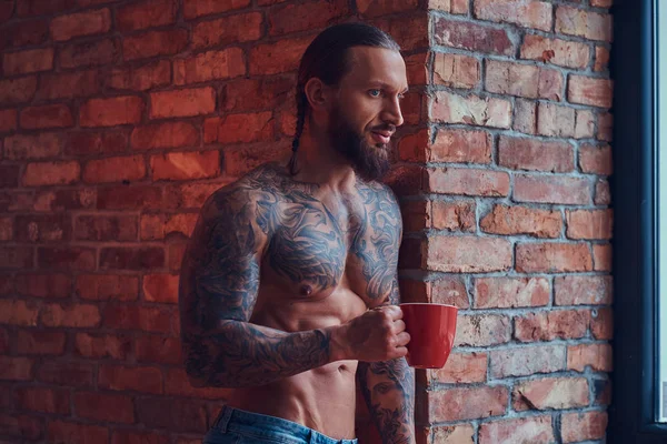 Portrait of a tattooed shirtless male with a stylish haircut and beard, drinks morning coffee, leaning against a brick wall in a room.