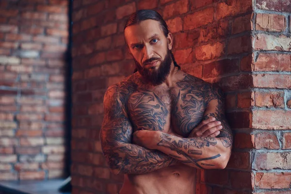 Handsome tattooed shirtless male with a stylish haircut and beard, standing with crossed arms, leaning against a brick wall in a room with a loft interior.