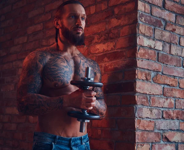 Handsome tattooed shirtless male with a stylish haircut and beard, doing exercise with a dumbbell, standing against a brick wall in a room with a loft interior.