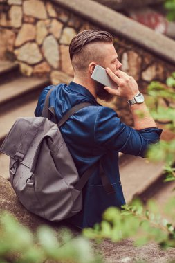 Bir saç kesimi ile sakallı bir erkek portresi arkadan görünüm ile telefonla konuşurken bir parkta oturan bir sırt çantası, rahat kıyafetler giymiş.