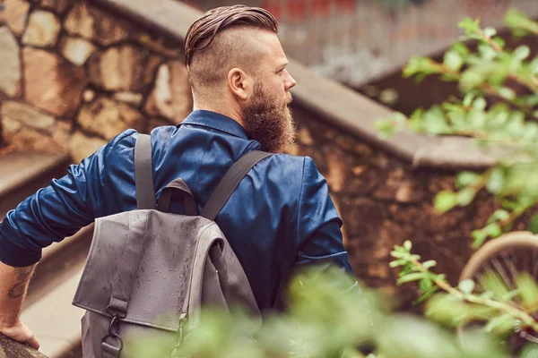 Back view portrait of a bearded male with a haircut dressed in casual clothes with a backpack, sitting in a park