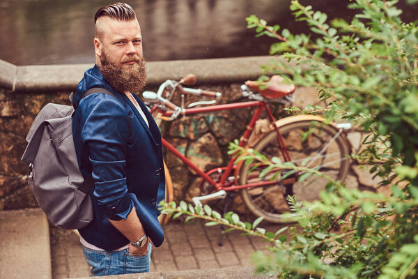 Portrait of a bearded male with a haircut dressed in casual clothes with a backpack, standing in a city park.