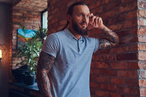 Um belo homem tatuado com um corte de cabelo elegante e barba, em uma camiseta cinza, de pé encostado a uma parede de tijolos em uma sala com um interior loft . — Fotografia de Stock