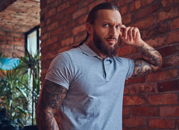 Un guapo hombre tatuado con un corte de pelo elegante y barba, en una camiseta gris, de pie apoyado contra una pared de ladrillo en una habitación con un interior loft . — Foto de Stock