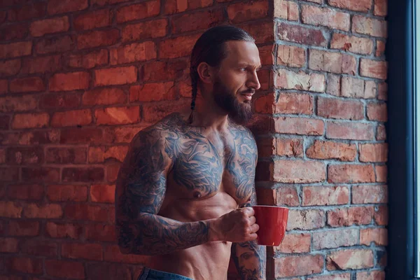 Retrato de un hombre sin camisa tatuado con un corte de pelo elegante y barba, bebe café de la mañana, apoyado contra una pared de ladrillo en una habitación con un interior loft . — Foto de Stock