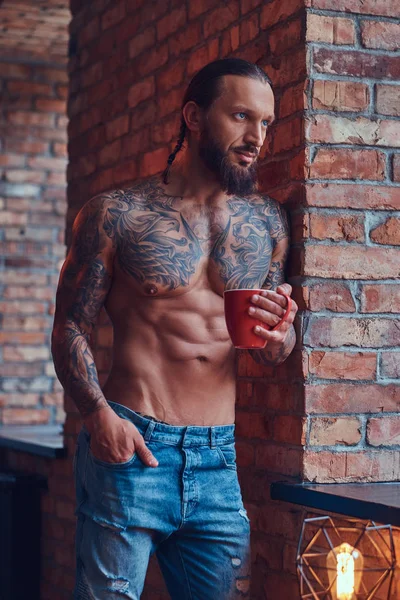 Portret van een man tattoed shirtless met een stijlvol kapsel en baard, drinkt koffie in de ochtend, leunend tegen een muur in een kamer met een loft interieur. — Stockfoto