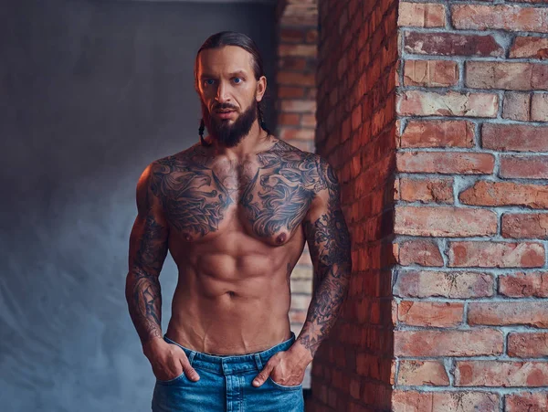 A handsome tattoed shirtless male with a stylish haircut and beard, standing leaning against a brick wall in a kitchen.