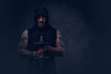 Bir kas sakallı tattoed erkek portresi karanlık bir arka plan üzerinde siyah spor bir kukuleta ile giyiyor, bir dumbbell ile egzersiz yaparak izole.