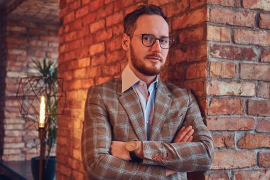 Pazen elbise giyip gözlük çatı iç ile bir odada bir tuğla duvara yaslanmış çapraz kollu şık adam.