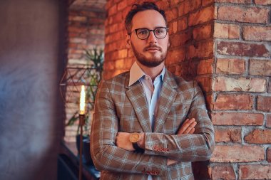 Pazen elbise giyip gözlük çatı iç ile bir odada bir tuğla duvara yaslanmış çapraz kollu şık adam.