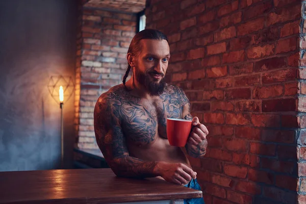 En stilig skäggiga tatueras hane med en snygg frisyr och muskulös kropp, drycker kaffe, lutande på en tabell. — Stockfoto