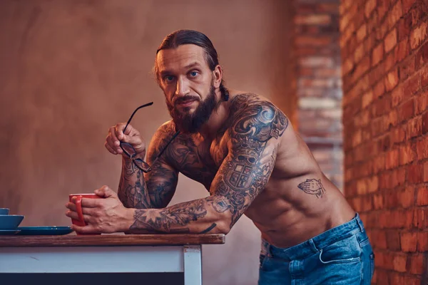 Een knappe bebaarde tattoed man met een stijlvol kapsel en gespierd lichaam, drinken koffie, leunend op een tabel. — Stockfoto
