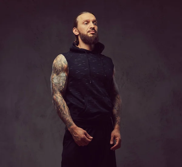 Retrato de un macho musculoso barbudo tatuado con un corte de pelo elegante con ropa deportiva negra, aislado sobre un fondo oscuro . — Foto de Stock