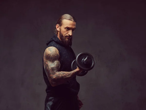 Портрет мускулистого бородатого татуированного мужчины со стильной стрижкой в черной спортивной одежде, делающего упражнения с гантелями, изолированного на темном фоне . — стоковое фото