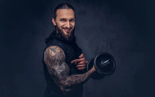 Portret van een man met een stijlvol kapsel dragen zwarte sportkleding, oefening met een halter, geïsoleerd op een donkere achtergrond doet gespierde bebaarde tattoed. — Stockfoto