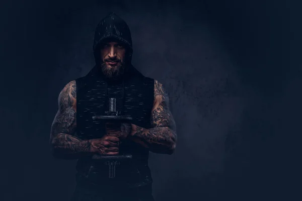 Ritratto di un maschio muscoloso barbuto tatuato che indossa abbigliamento sportivo nero con cappuccio, facendo esercizio con un manubrio, isolato su uno sfondo scuro . — Foto Stock