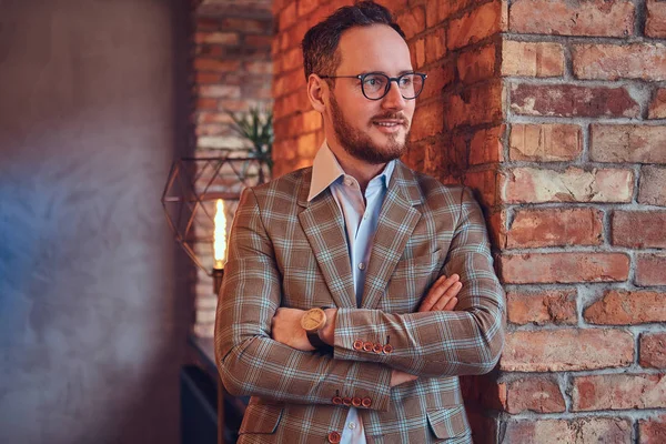 Stilvoller Mann im Flanellanzug und Brille mit verschränkten Armen, der sich in einem Raum mit Dachboden an eine Ziegelwand lehnt. — Stockfoto