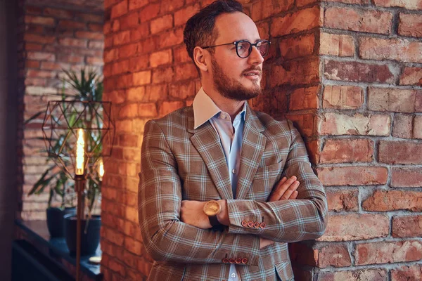 Elegante hombre con traje de franela y gafas con brazos cruzados apoyados en una pared de ladrillo en una habitación con interior tipo loft . — Foto de Stock