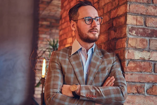 Stilvoller Mann im Flanellanzug und Brille mit verschränkten Armen, der sich in einem Raum mit Dachboden an eine Ziegelwand lehnt. — Stockfoto