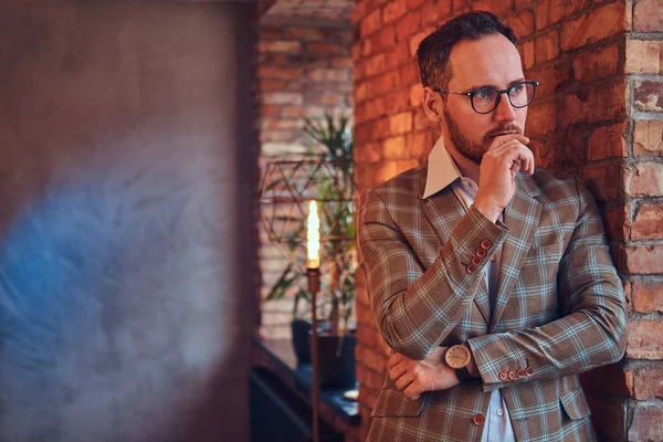 Porträt eines stilvollen Mannes in Flanellanzug und Brille, der sich in einem Raum mit Loft-Interieur an eine Ziegelwand lehnt. — Stockfoto