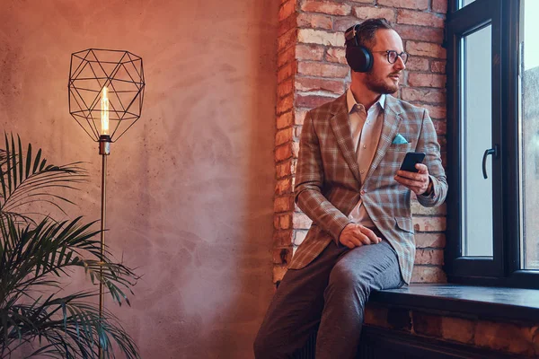Retrato de un hombre con estilo en un traje de franela y gafas escuchando música con el teléfono y los auriculares sentados en un alféizar de la ventana en una habitación con interior loft . — Foto de Stock