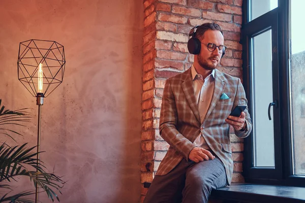 Porträt eines stilvollen Mannes in Flanellanzug und Brille, der mit Telefon und Kopfhörer Musik hört, auf einem Fensterbrett in einem Raum mit Loft-Interieur sitzt. — Stockfoto