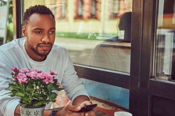 Un hombre barbudo afroamericano de moda sentado cerca de una cafetería con una taza de café, usando un teléfono inteligente . —  Fotos de Stock