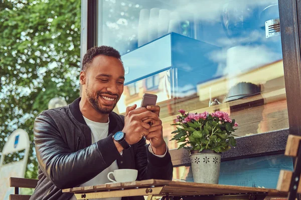 En fashionabla afroamerikanska skäggiga manliga sitter nära ett kafé med en kopp kaffe, med en smartphone. — Stockfoto