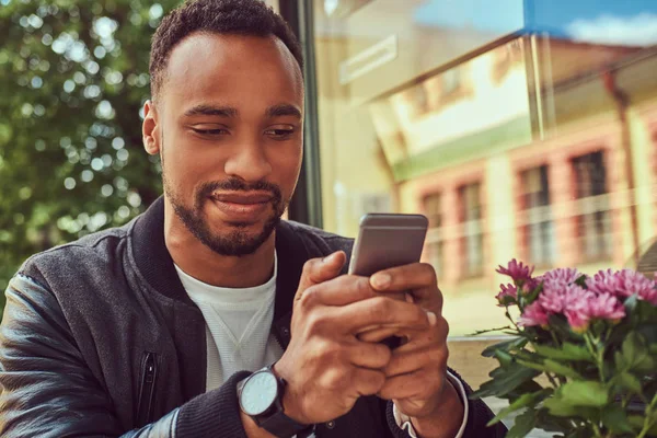 En fashionabla afroamerikanska skäggiga manliga sitter nära ett kafé med en kopp kaffe, med en smartphone. — Stockfoto