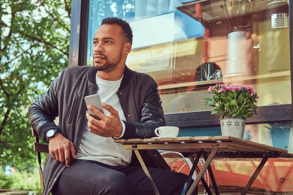 Un hombre barbudo afroamericano de moda sentado cerca de una cafetería con una taza de café, usando un teléfono inteligente . —  Fotos de Stock