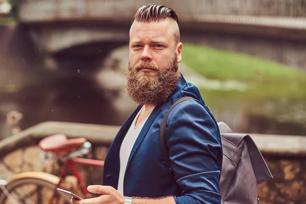 Retrato de un hombre barbudo con un corte de pelo vestido con ropa casual con una mochila, de pie en un parque, usando un teléfono inteligente . — Foto de Stock