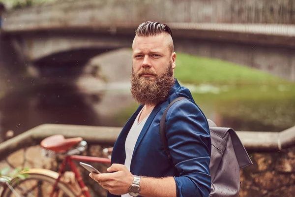 Retrato de un hombre barbudo con un corte de pelo vestido con ropa casual con una mochila, de pie en un parque, usando un teléfono inteligente . —  Fotos de Stock