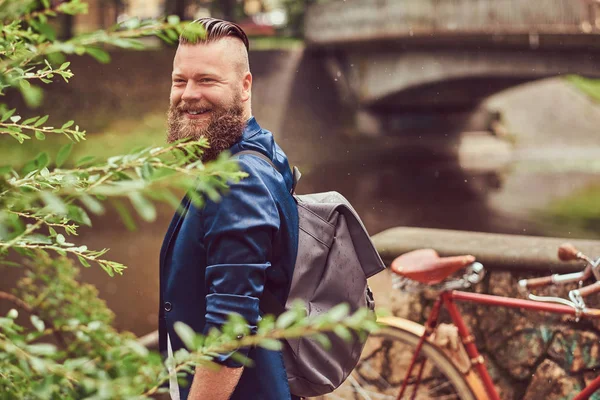 Retrato de un hombre barbudo con un corte de pelo vestido con ropa casual con una mochila, de pie en un parque . — Foto de Stock