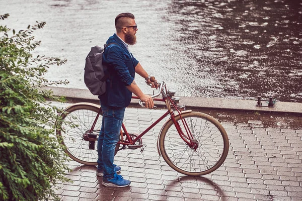 Um homem barbudo com um corte de cabelo vestido com roupas casuais e óculos de sol, de pé na chuva, em um parque . — Fotografia de Stock